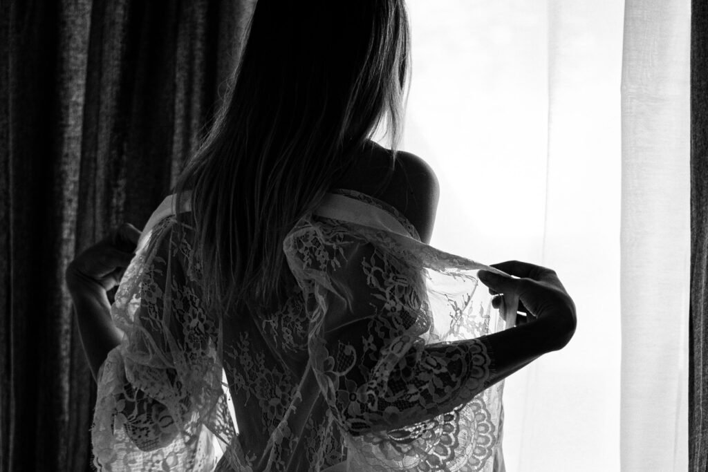 Girl standing in front of a window with her back towards us, letting her white lace night robe slip from her shoulders
