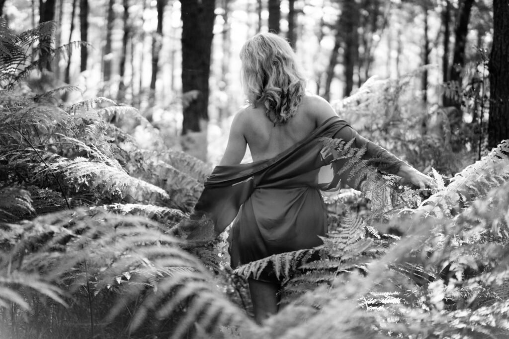 Woman with blond hair wearing a dark night robe, walking in a forest between large ferns with her back towards us. Her night robe hanging down her shoulders.