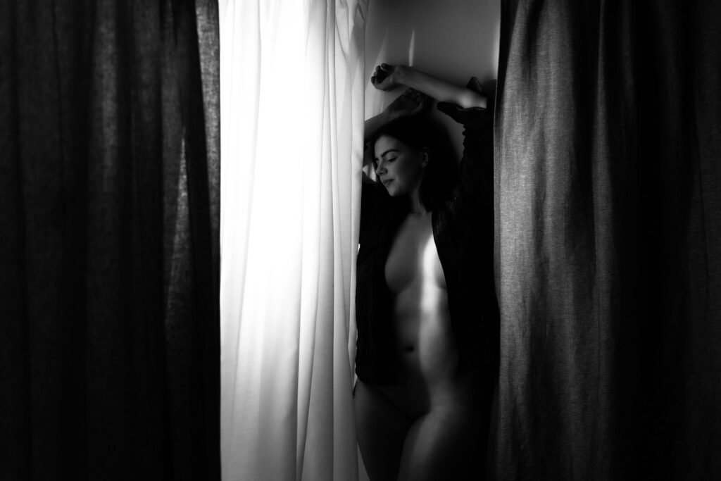 Naked girl wearing a black open button shirt leaning with her back against the wall next to a window with her arms above her head.