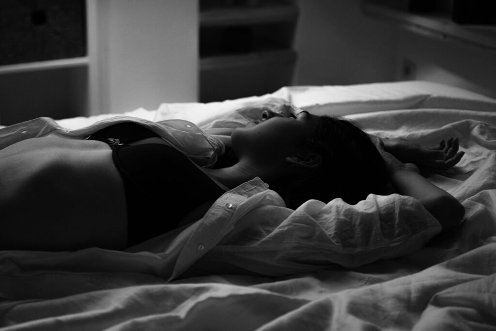 Girl laying on her back on the bed with her arms above her head, her button shirt casually unbuttoned revealing her black bra.